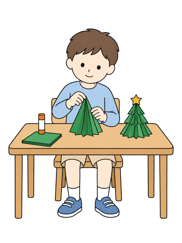 男の子が机で緑の紙を折り、立体的なクリスマスツリーを作っている。そばに完成した小さな紙ツリーがある。